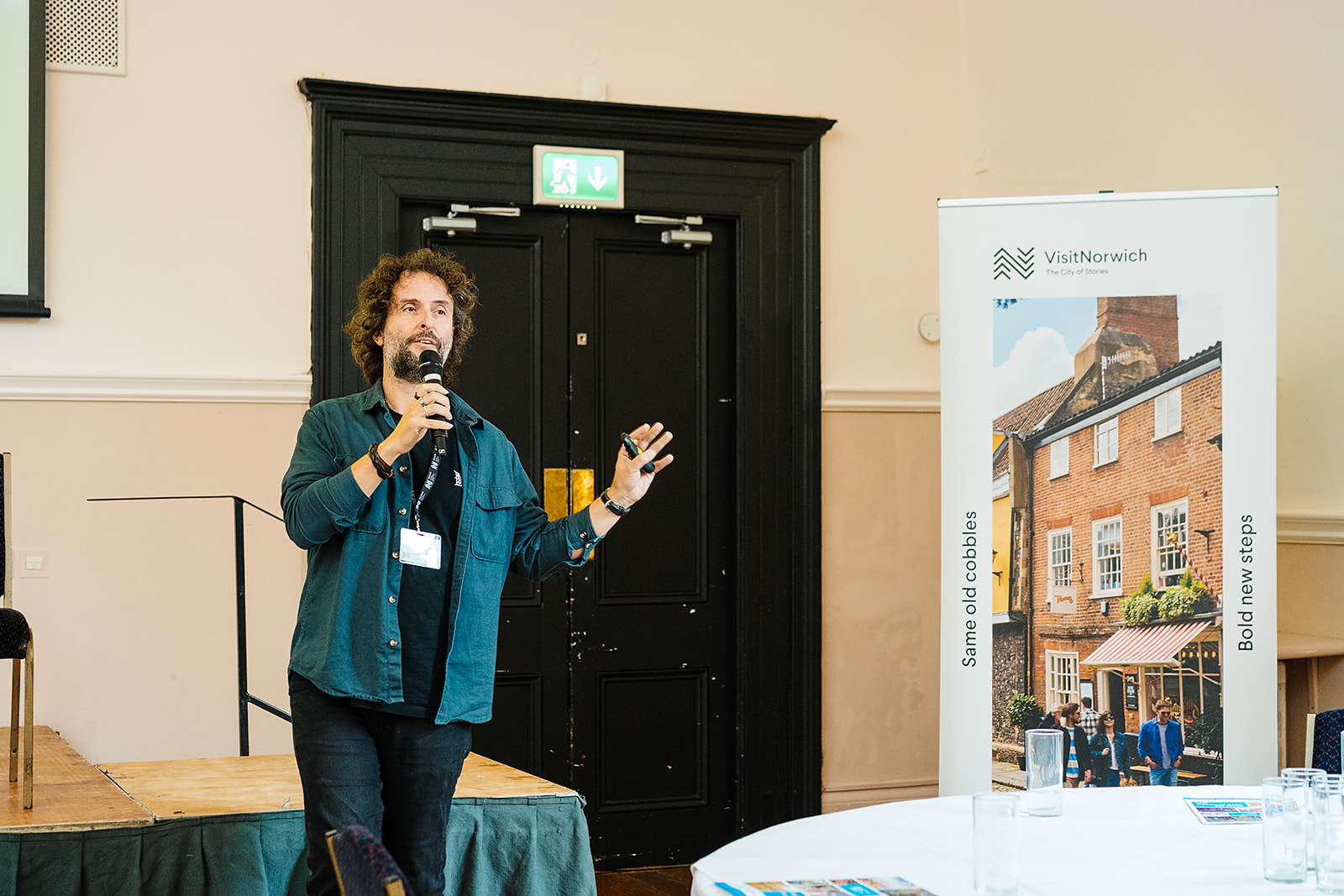 A photo of a white man with dark curly hair and a beard. He is inside what looks like an event space, holding a microphone and talking. Next to him is a poster for the Visit Norwich campaign. It shows an old building, shops, people walking in the sunshine. The copy on the banner reads: Same old cobbles, bold new steps.