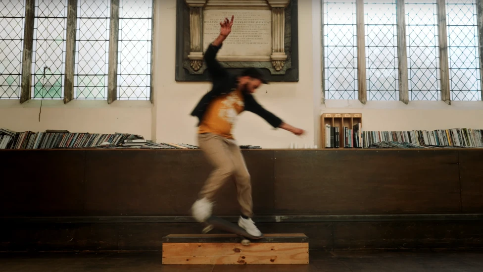 A photo taken inside a church. There are two large windows, an old memorial on the wall, and books/magazines lining the shelf infant of the windows. In the centre is a man on a skateboard. He's wearing khaki trousers, a yellow t-shirt, and dark over shirt. He's blurred because he's in motion.