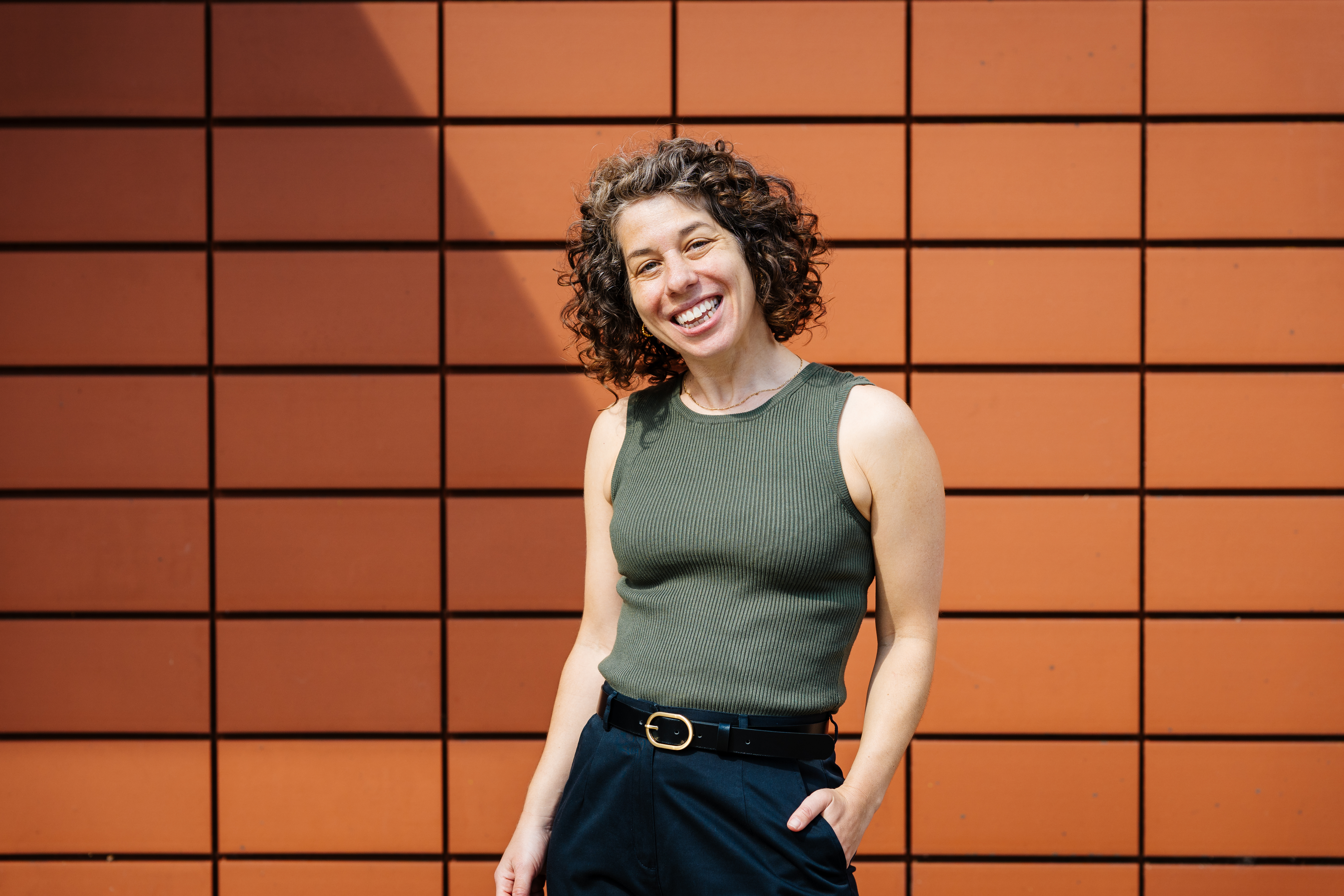 A photo of Sarah Herman, a white woman with dark, short curly hair. Sarah is smiling. She's wearing a green top and black trousers and is stood in front of an orange tiled wall.